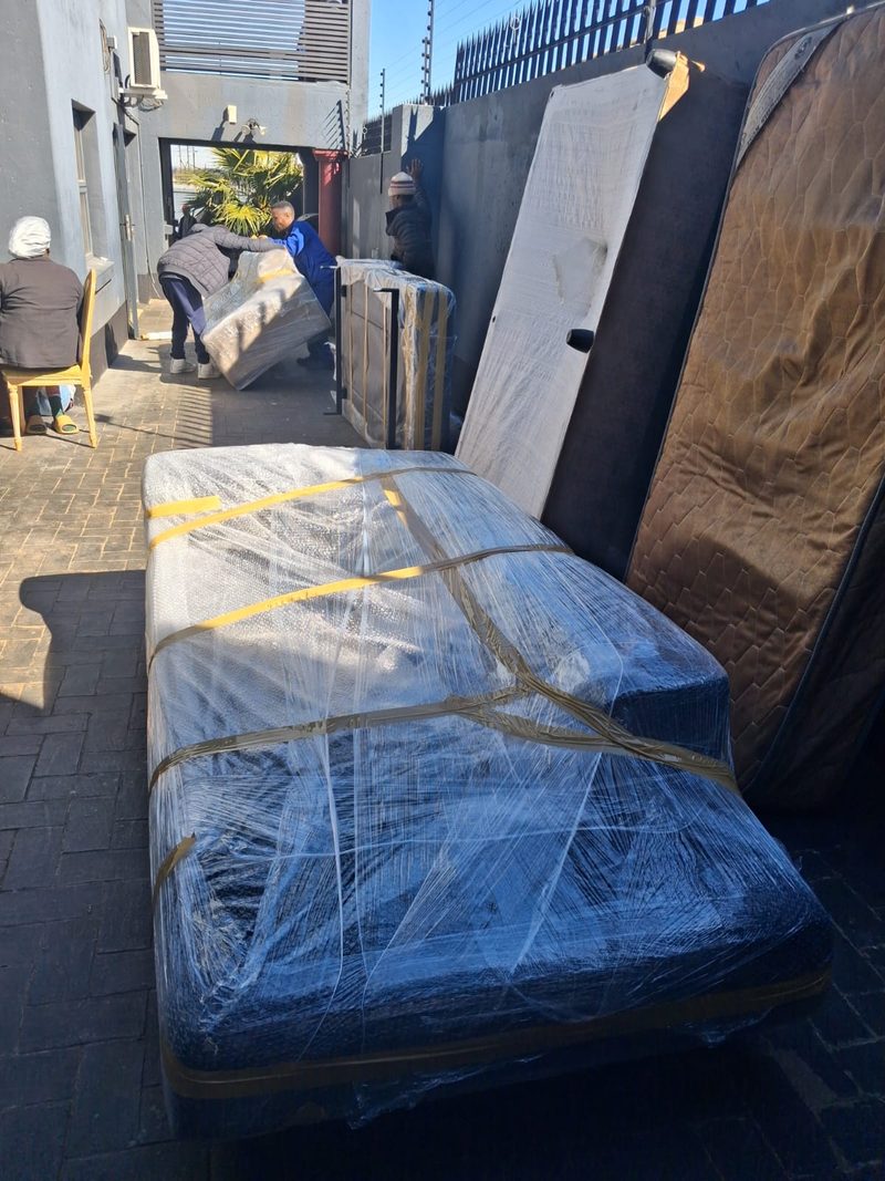 A bed base sealed in plastic and strapped with tape standing on paving, with mattresses and a door leaning against the wall behind it.