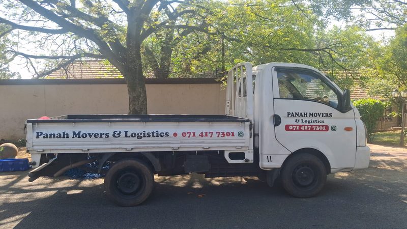 A white Panah Movers bakkie parked in the shade of a tree, its drop-side panel lettered with the company name and phone number.