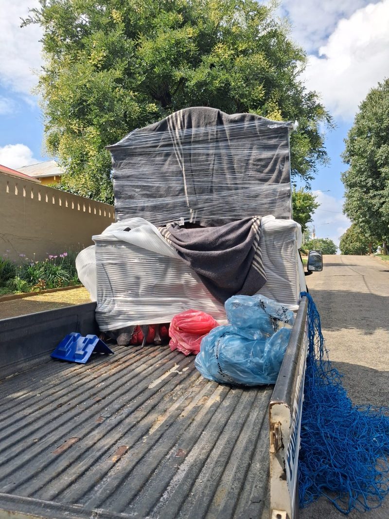 The open back of a bakkie carrying a wrapped headboard and mattress under blankets, with cargo netting folded on the tailgate.