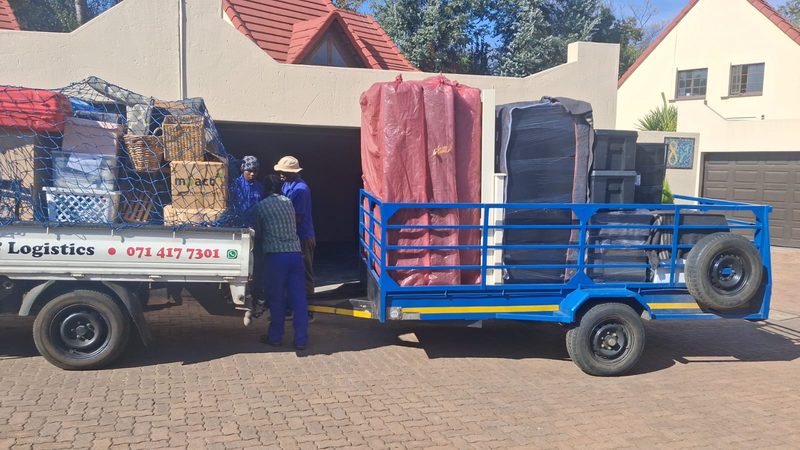 A loaded Panah bakkie and blue trailer outside a house, stacked with wrapped mattresses, baskets and cartons held down under cargo netting.