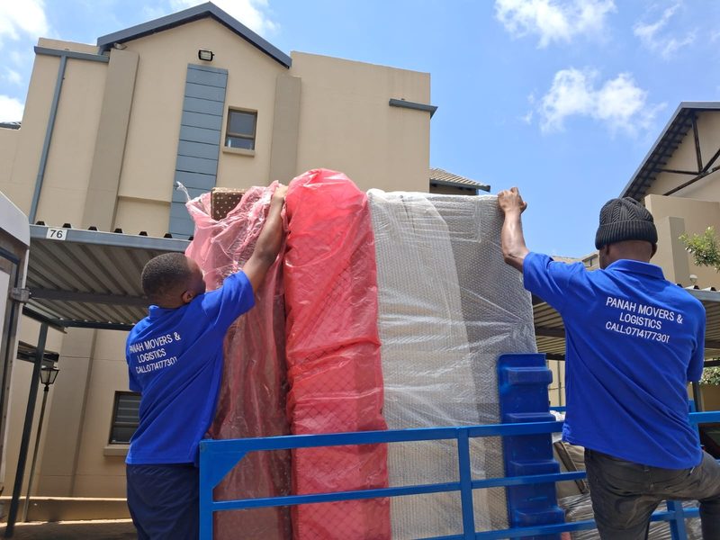 Two crew in branded blue shirts lifting a wrapped mattress and bed base onto a blue trailer outside a townhouse.