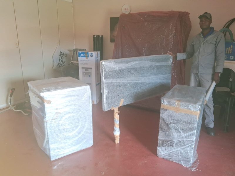 A crew member standing beside a washing machine, a headboard and a cabinet, each wrapped in bubble wrap and taped, in an empty room.