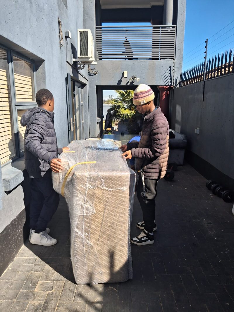 Two crew members wrapping a couch in bubble wrap and yellow tape on a paved driveway.