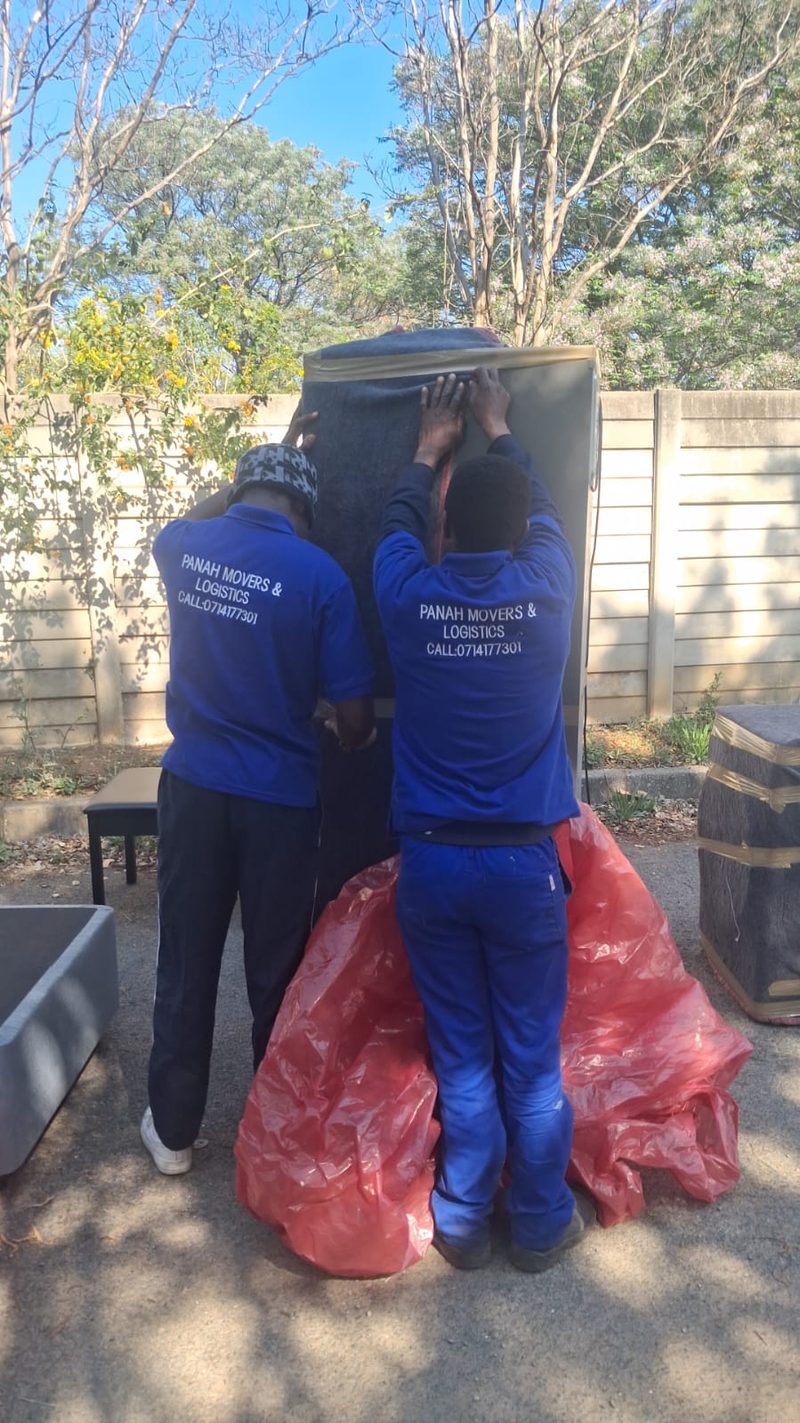 Two crew members in branded shirts covering a fridge with a blanket and taping it closed in a yard.